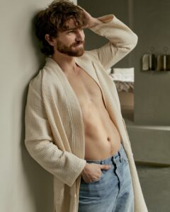 Een man met golvend bruin haar en een baard draagt de Vandyck Biarritz New Badjas in de kleur cream tan XS over een blauwe spijkerbroek en poseert nonchalant tegen een muur.