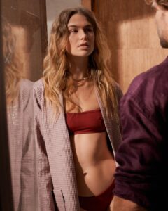 Een vrouw draagt een Vandyck Biarritz New Badjas in de kleur mauve, maat XL, en kijkt aandachtig naar een man; haar spiegelbeeld is in het glas te zien.