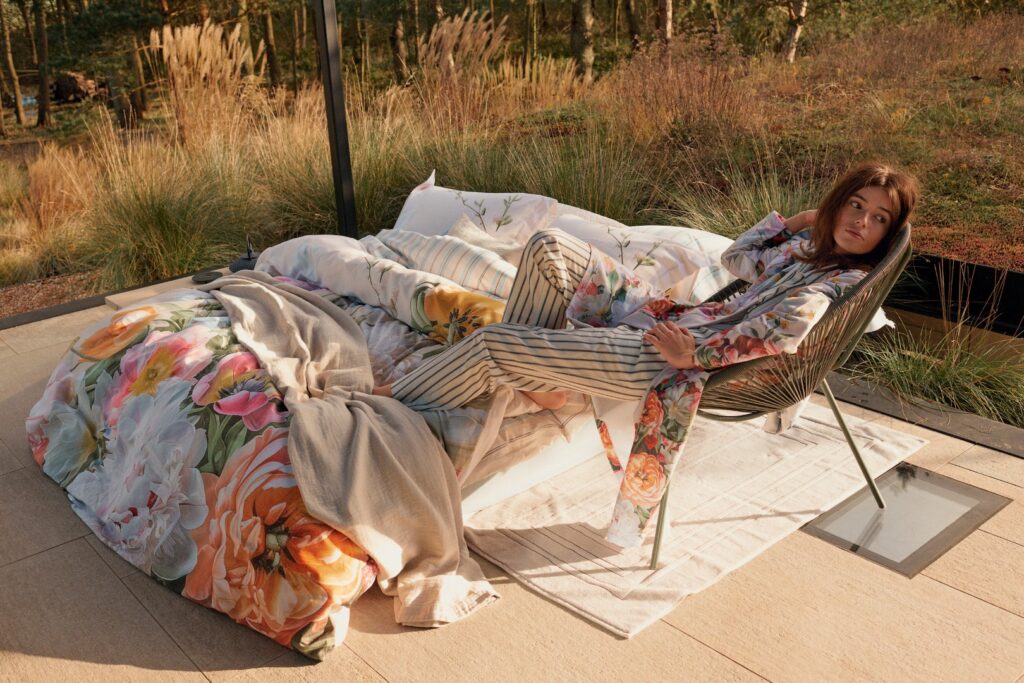 Een vrouw in een Vandyck Fleur-badjas in de kleur lavender blue L zit ontspannen op een stoel naast een bed met een vrolijk bloemetjesdekbed op het terras.