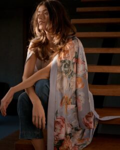 Een vrouw met lang bruin haar zit ontspannen op houten traptreden, gekleed in een lavendelblauwe Vandyck Fleur-badjas over lichte kleding.
