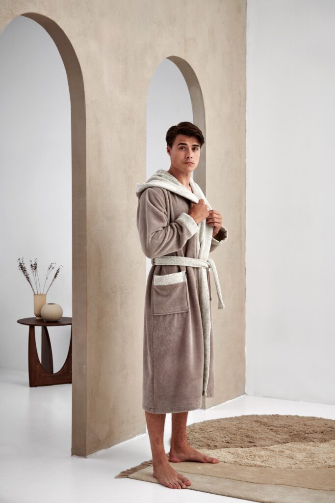 Een jonge man staat op blote voeten op een beige vloerkleed en draagt de Vandyck Georgia-badjas in maat XL voor een lichte muur met bogen.