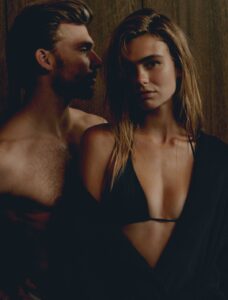 Een vrouw in een zwarte bikinitop en een donkerblauwe Vandyck Grace Badjas S staat voor een shirtloze man tegen een donkere houten achtergrond.