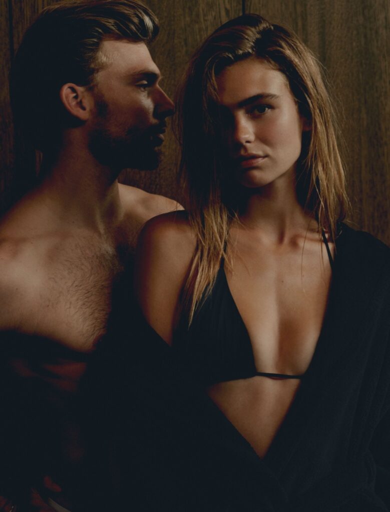 Tegen een donkere houten achtergrond staan een man zonder shirt en een vrouw in een zwarte bikini, met de Vandyck Grace-badjas XL ernaast.