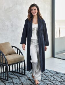 Een vrouw loopt glimlachend op blote voeten door het huis langs een raam, gekleed in een donkerblauwe Vandyck Grace-badjas en een witte satijnen pyjama, naast een zwarte stoel met beige kussens.
