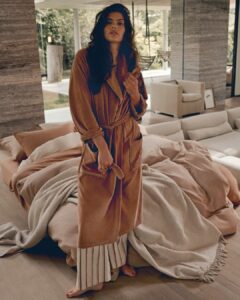 Een vrouw op blote voeten met lang donker haar staat naast een onopgemaakt bed in een zonnige, moderne kamer. Ze draagt de Vandyck Grace-badjas in de kleur fudge S en een gestreepte broek, omringd door zachte accessoires en beige dekens.