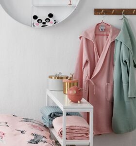 Op de plank liggen handdoeken, een roze knuffel en gouden doosjes naast twee badjassen, waaronder de lichtroze Vandyck Kids-badjas voor meisjes. Boven hangt een ronde panda-plank, vlak bij een bed met een roze dekbed.