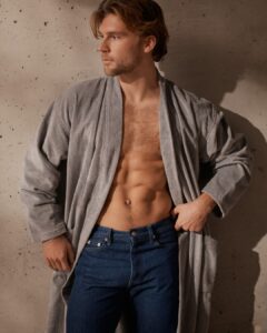 Een man met lichtbruin haar draagt de grijze Vandyck Kyoto-badjas losjes over zijn blote bovenlichaam en een blauwe spijkerbroek, terwijl hij opzij kijkt.