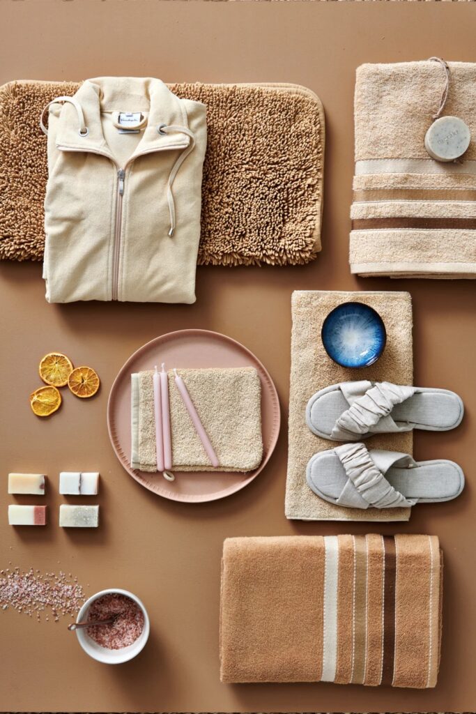 De Vandyck Prestige strandhanddoekenset in beige en zandkleur ligt naast slippers, kaarsen, zeep, chocolademelk, een blauw schaaltje en schijfjes sinaasappel.