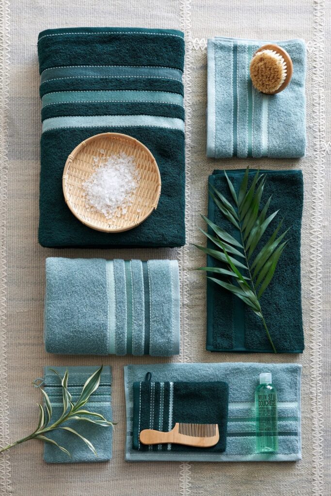 De Vandyck Prestige-handdoekenset in donkergroen vormt samen met een houten borstel, een bamboe schaaltje, een flesje en groene takjes een harmonieus geheel op een beige achtergrond.