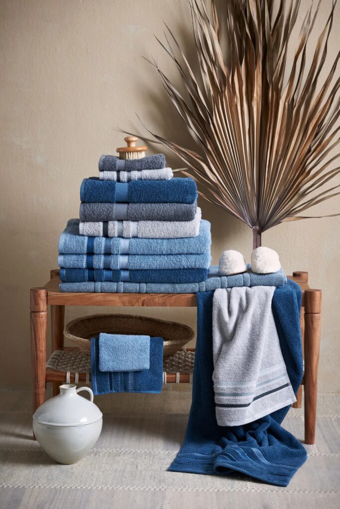 Op een houten bank liggen de Vandyck Prestige-wastafelset in Dusty Blue, samen met een lichaamsborstel, slippers, een kruik en een palmblad.