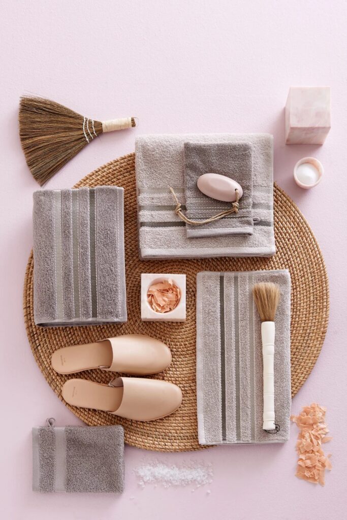 De Vandyck Prestige-handdoekenset Hazel vormt samen met beige badslippers, een strooien mat, zeep en accessoires een sfeervol geheel.