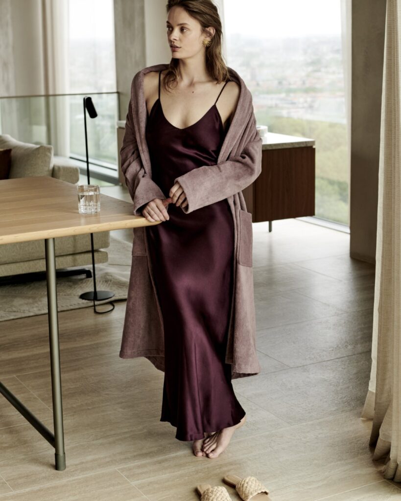 Een vrouw in een kastanjebruine satijnen slipdress met de Vandyck Prestige uni mauve XL-badjas leunt op blote voeten tegen een houten tafel met een glas water, met gevlochten slippers en een groot raam op de achtergrond.
