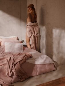 Een vrouw in een bruine outfit en met een pet op staat naast een onopgemaakt bed met Vandyck Prestige uni sepia pink S-beddengoed in een zacht verlichte, minimalistische kamer met betonnen muren.
