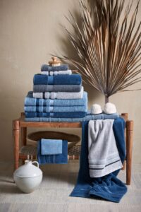 De Vandyck Prestige-douchehanddoekenset in Vintage Blue ligt stijlvol opgestapeld op een houten bankje, samen met badaccessoires en een palmblad.