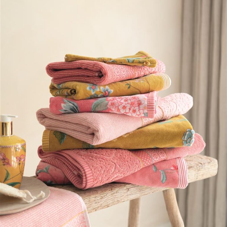 Op een houten bankje liggen netjes opgevouwen roze en okergele handdoeken met bloemenprint uit de Pip Studio-verhalencollectie, naast een zeepdispenser.