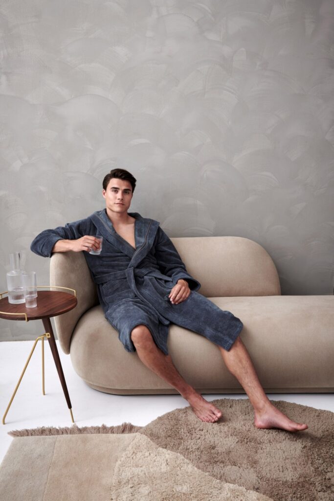 Een man zit ontspannen op een beige bank in een Vandyck Taylor Oceanview M-badjas met een glas water, naast hem staat een tafeltje.