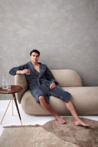 Op een beige bank zit een ontspannen man in een Vandyck Taylor-badjas in de kleur Oceanview Blue, met een glas water naast zich op tafel.