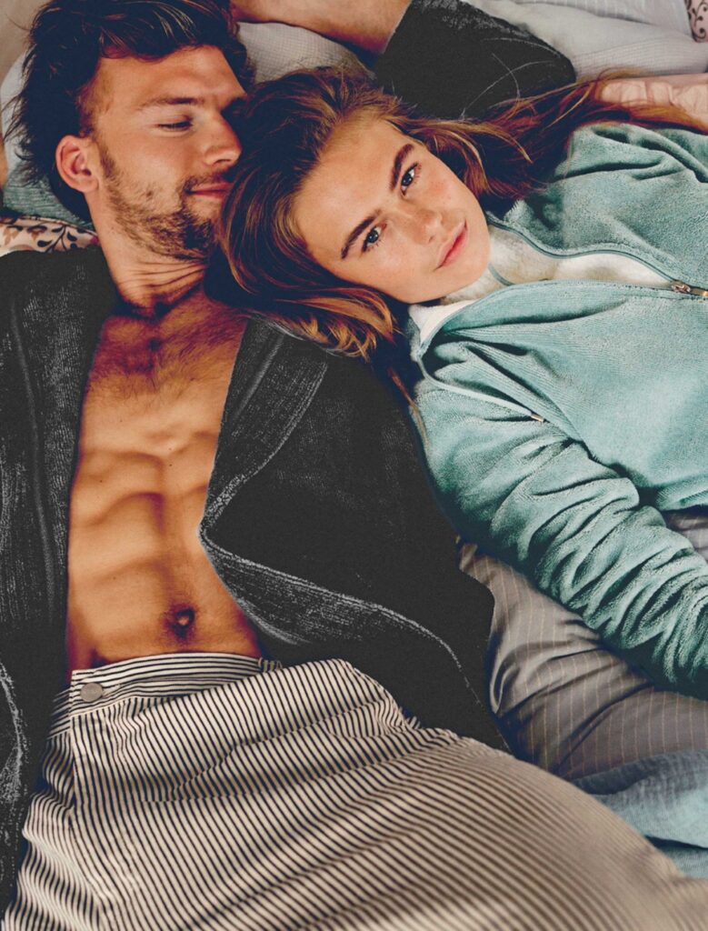 Een jonge man in een zwarte kamerjas en een vrouw in een lichtblauwe trui met rits zitten samen ontspannen op bed en stralen rust uit in deze Vandyck Taylor Oceanview M-sfeer.