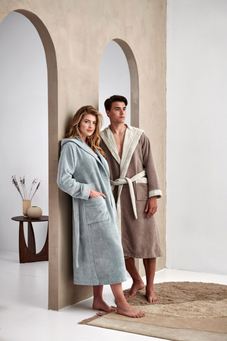 Een vrouw en een man dragen parelblauwe Vandyck Taylor-badjassen in maat M en staan samen bij een boogdeur in een rustige, minimalistische kamer.