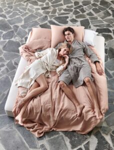 Een man en een vrouw in badjassen liggen te ontspannen op een bed met Vandyck Toledo Steelgrey M-beddengoed op een stenen vloer.