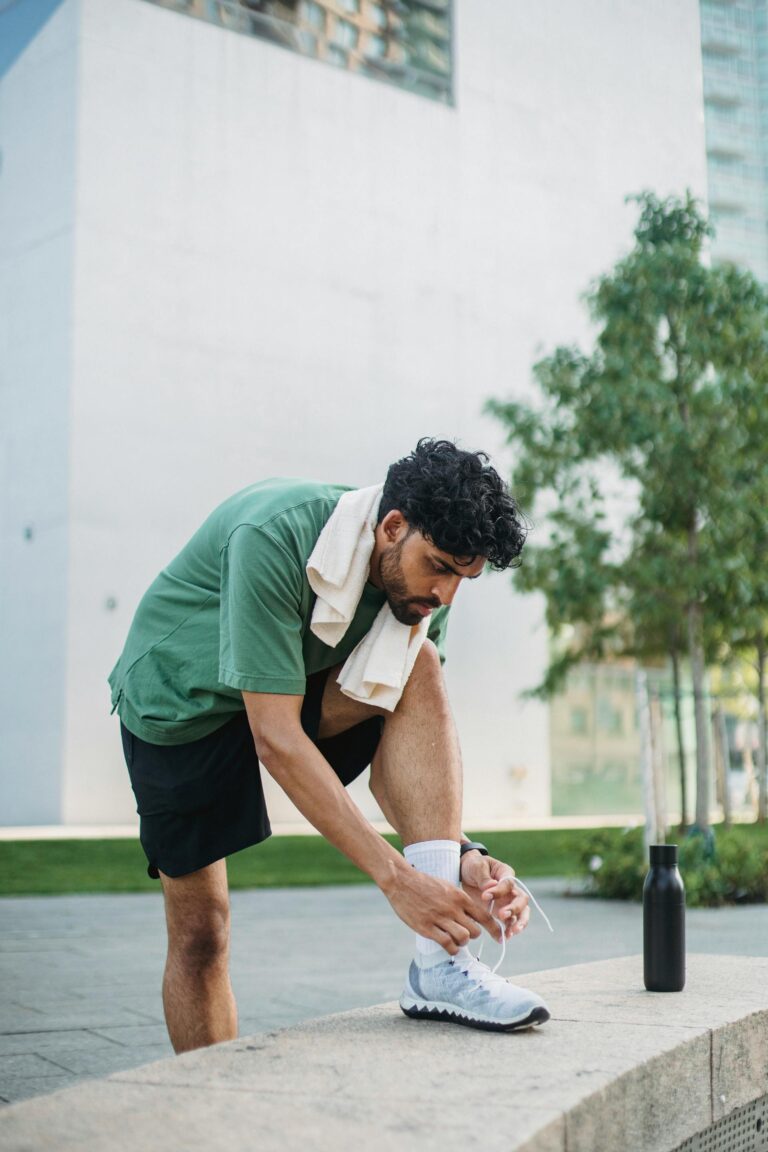 Een man in een groen shirt en een zwarte korte broek knoopt buiten op een richel zijn sportschoenen vast; hij heeft een sporthanddoek om zijn nek en er staat een waterfles naast hem.