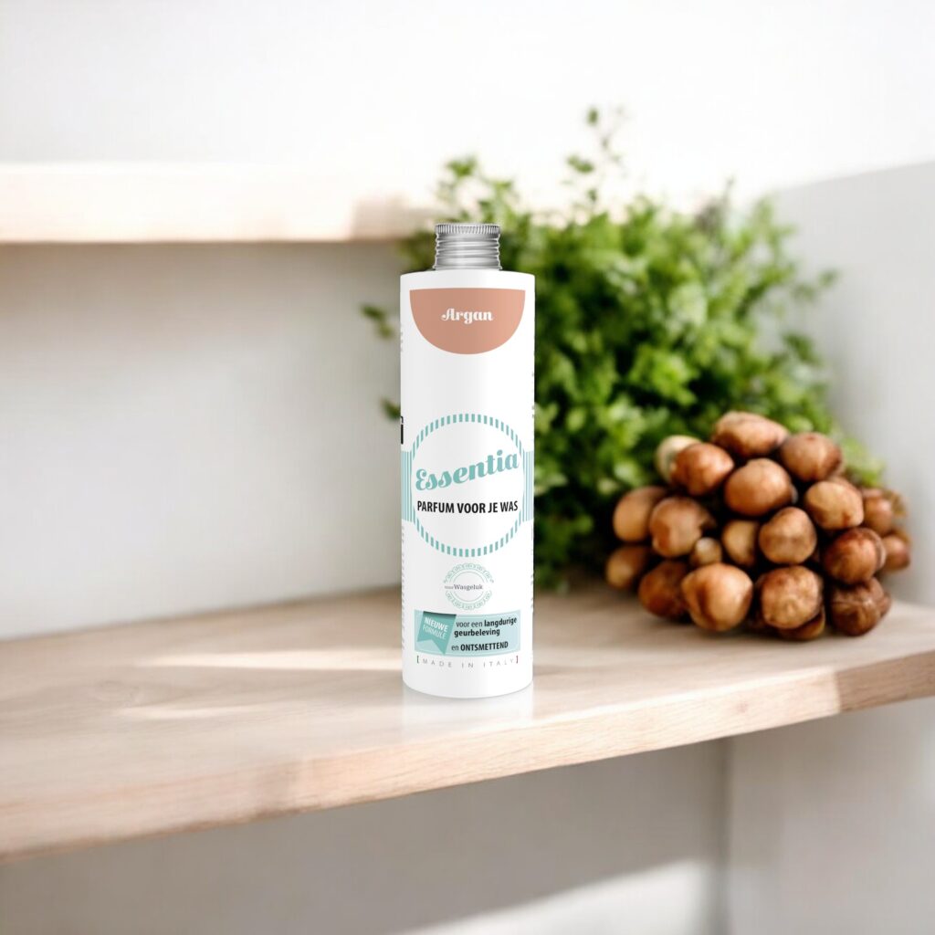 Op een houten plank staat Essentia Wasparfum Argan naast argannoten, met op de achtergrond vaag een groene plant te zien.