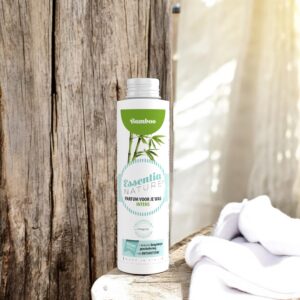 Op een rustiek houten oppervlak staat een fles Essentia Wasparfum Bamboo naast opgevouwen witte handdoeken in het zonlicht.