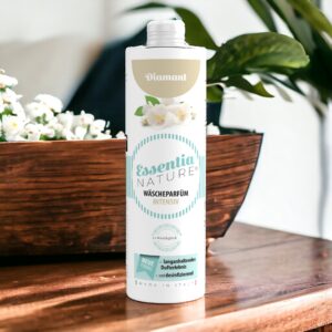 Essentia Wasparfum Diamant, met lichtblauwe details en bloemmotieven, staat op een houten voetstuk tussen witte bloemen en groene bladeren.