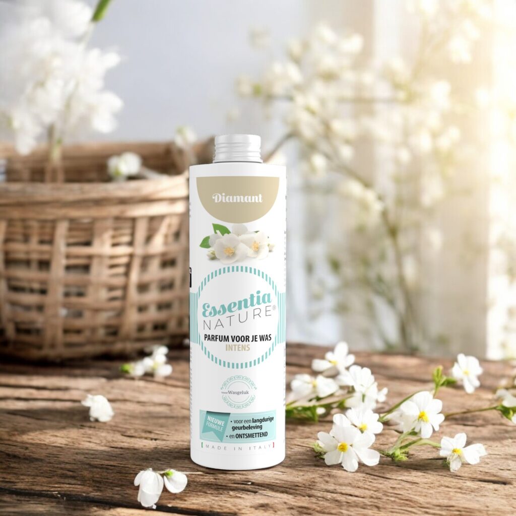 Op een houten ondergrond staat een flesje Essentia Wasparfum Diamant, omringd door witte bloemen en een sfeervolle mand met zachte bloesems.