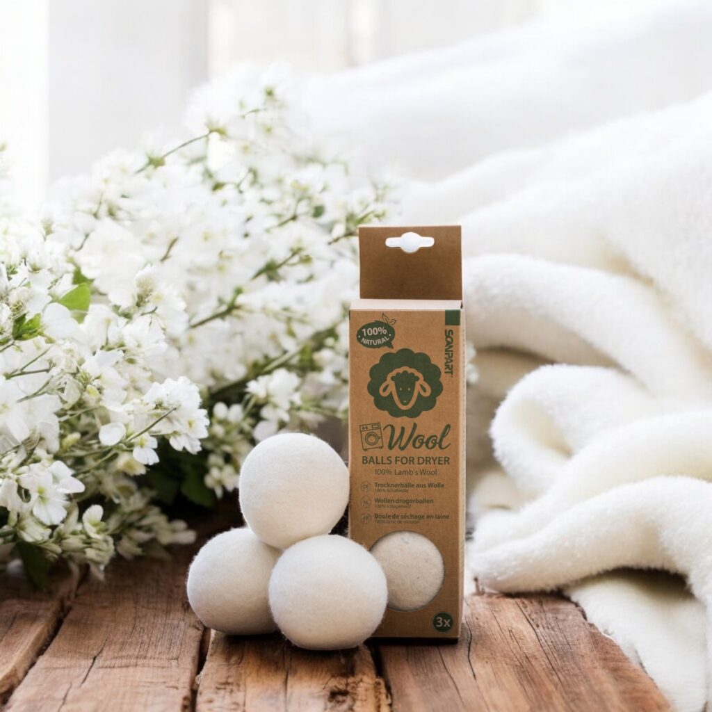 Op een houten ondergrond staat een doos Essentia Drogerballen met vier witte wollen drogerballen en sierlijke bloemen erbij.