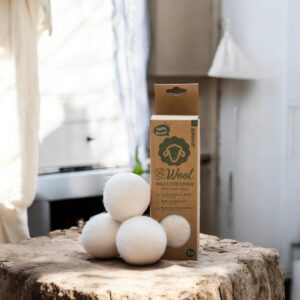 Op een houten ondergrond liggen Essentia-droogballen naast vier witte wollen droogballen, zacht verlicht door zonlicht.