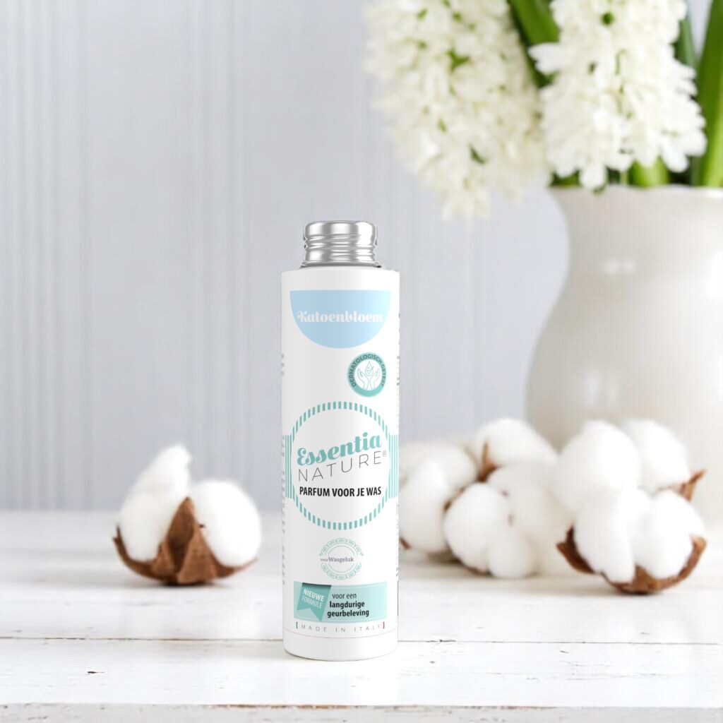 Een flesje Essentia Wasparfum Katoenbloem van 250 ml staat tussen wattenbolletjes, met daarachter een witte vaas met bloemen tegen een lichte houten wand.