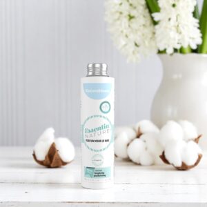 Een flesje Essentia Wasparfum Katoenbloem van 250 ml staat tussen wattenbolletjes, met daarachter een witte vaas met bloemen tegen een lichte houten wand.