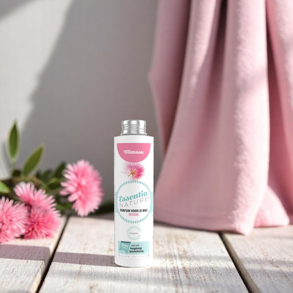Op een houten oppervlak staat een flesje Essentia Wasparfum Mimosa tussen roze bloemen en een zachtroze doek, wat zorgt voor een natuurlijke sfeer.