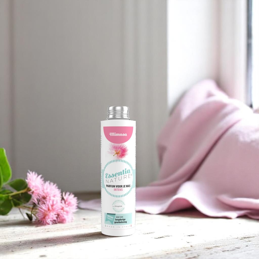 Op een houten oppervlak staat een fles Essentia Wasparfum Mimosa naast een roze bloem, met op de achtergrond een roze handdoek en een zacht verlicht raam.