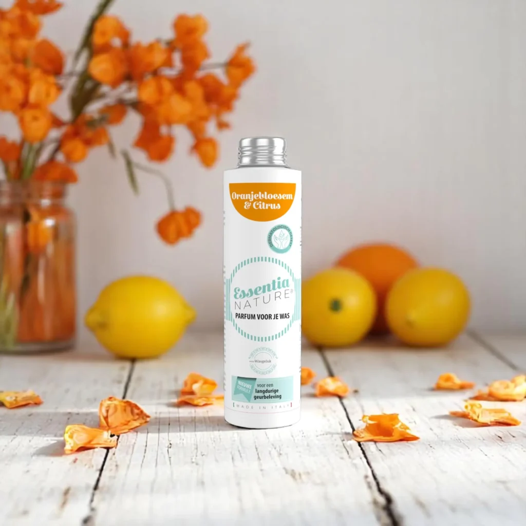 Op een houten ondergrond staat een fles Essentia Wasparfum Oranjebloesem en Citrus tussen oranjebloesems en citroenen, met op de achtergrond een vaas vol oranje bloemen.