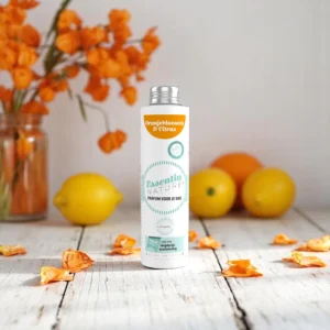 Op een houten ondergrond staat een fles Essentia Wasparfum Oranjebloesem en Citrus tussen oranjebloesems en citroenen, met op de achtergrond een vaas vol oranje bloemen.