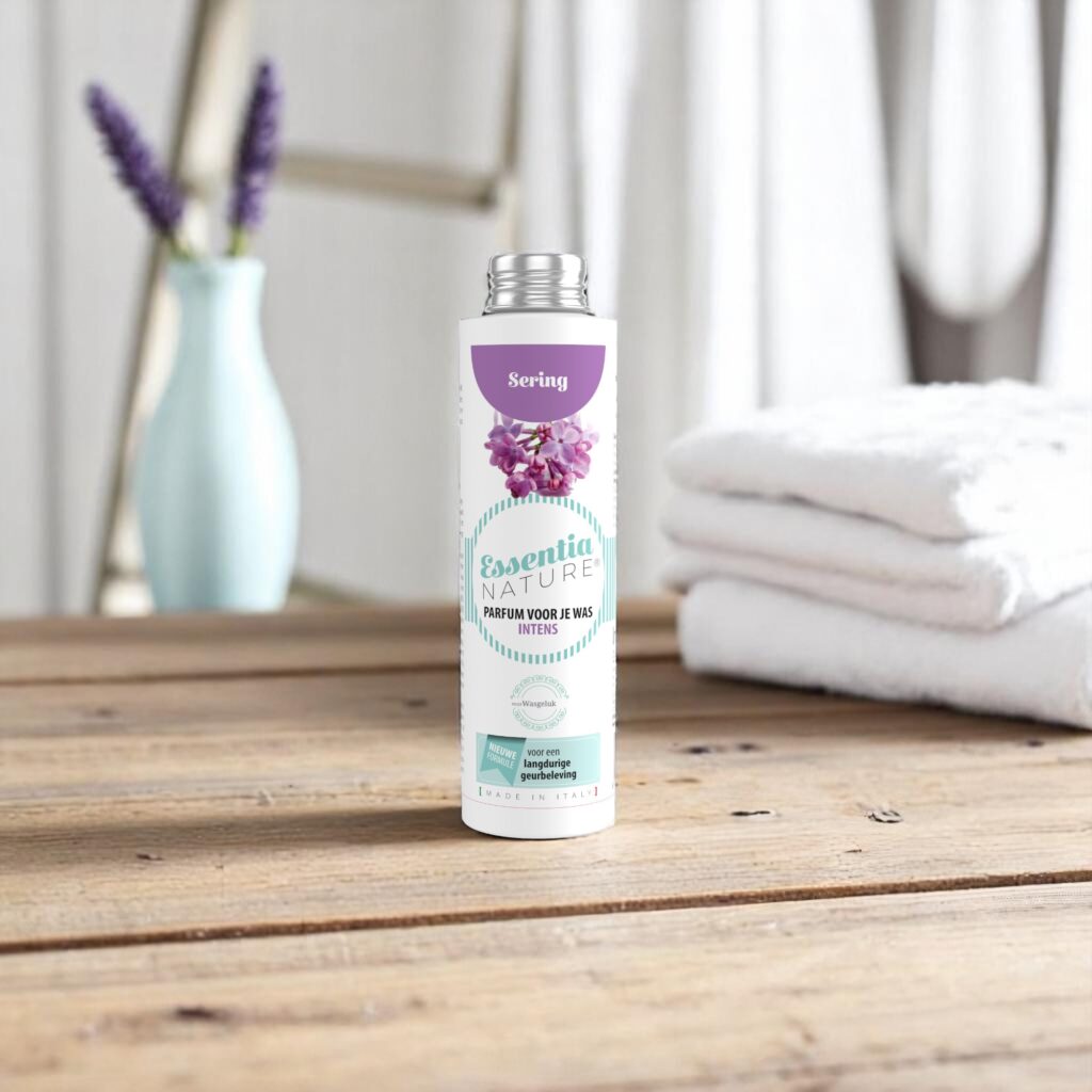 Op een houten tafel staat een flesje Essentia Wasparfum Sering, samen met een opgevouwen witte handdoek en paarse bloemen op de achtergrond.