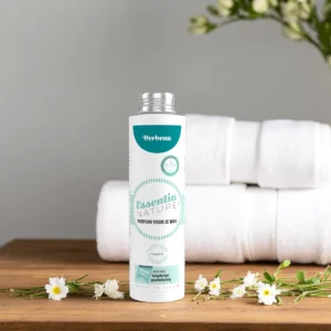 Essentia Waspafurm Verbena 250 ml staat op een houten oppervlak met kleine witte bloemen, opgerolde handdoeken en een grijze bloemenmuur.