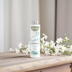Essentia Wasparfum Witte Musk 250 ml staat op een houten oppervlak naast kleine witte bloemen in een rustige, neutrale omgeving.