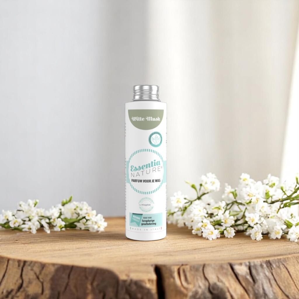 Op een houten oppervlak staat een fles Essentia Wasparfum Witte Musk, met witte bloemetjes en zacht licht dat door het gordijn schijnt.