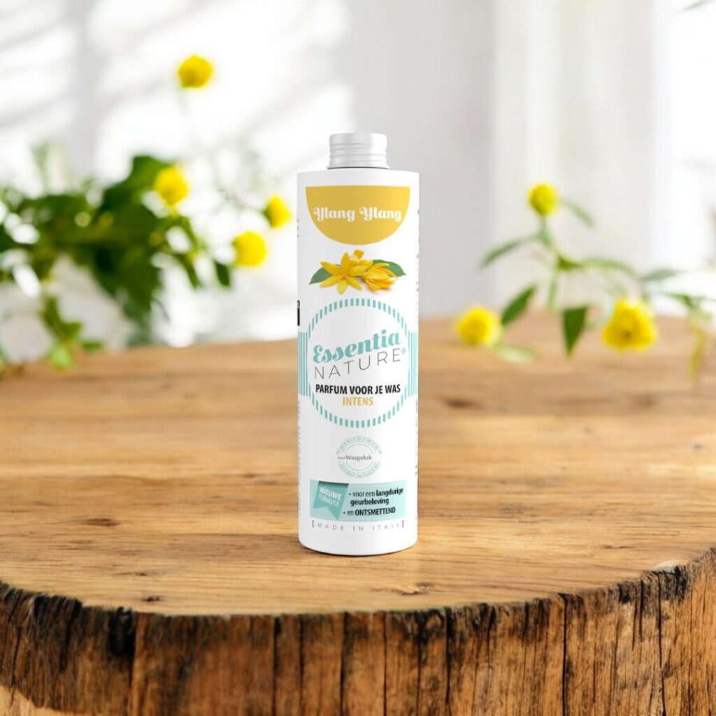 Op een houten ondergrond staat de Essentia Wasparfum Ylang Ylang-fles, met vaag gele bloemen en groen op de achtergrond.
