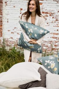 Een vrouw draagt een effen Vandyck Prestige-badjas in de kleur Cream tan (maat XS) en houdt een blauwgroen bloemenkussen vast, met witte en grijze kussens voor zich.