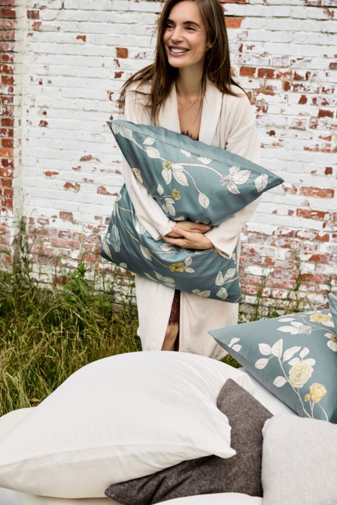 Een vrouw draagt een effen Vandyck Prestige-badjas in de kleur Cream tan (maat XS) en houdt een blauwgroen bloemenkussen vast, met witte en grijze kussens voor zich.