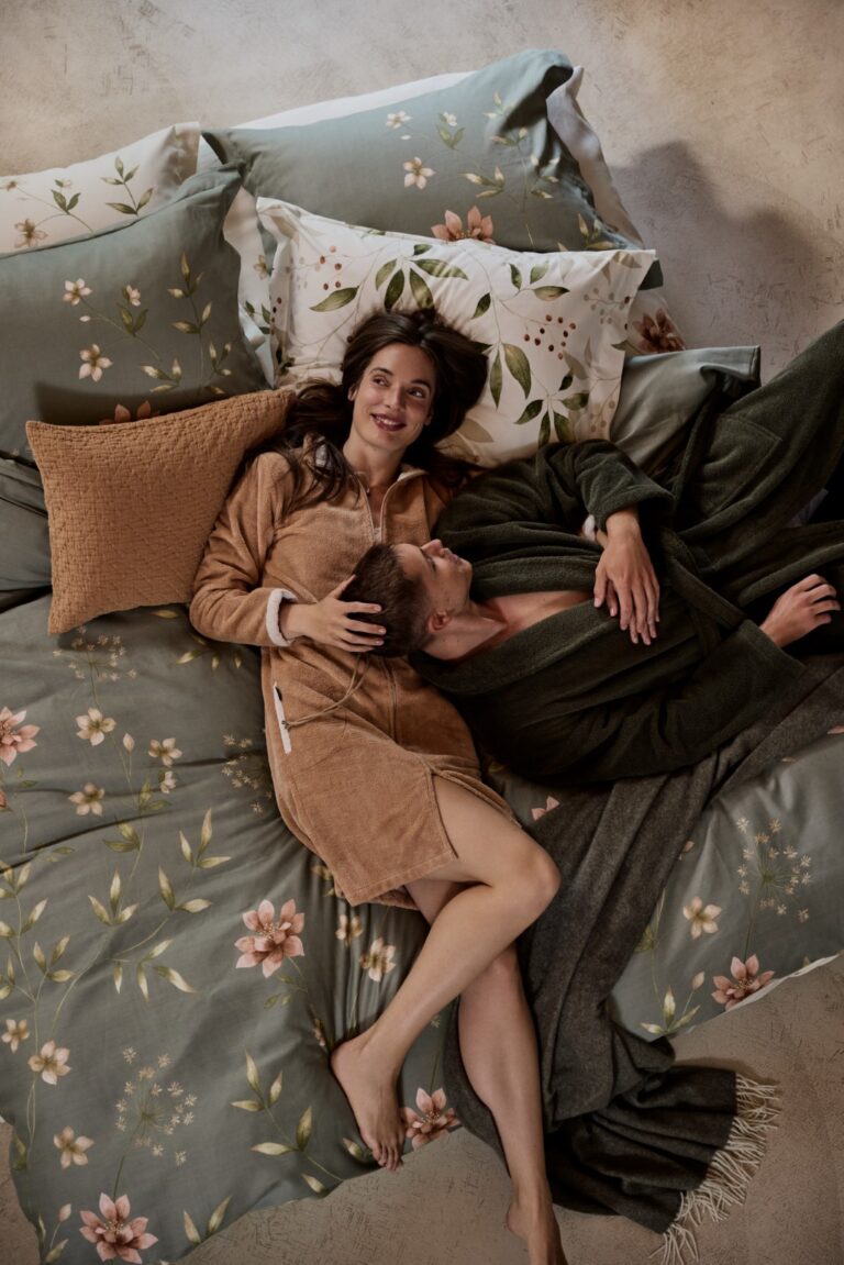 Een glimlachende vrouw in een beige badjas ligt achterover op groen en beige beddengoed met bloemenmotief, terwijl ze zachtjes een man in de Vandyck Ranger Badjas Thyme L in haar armen wiegt.