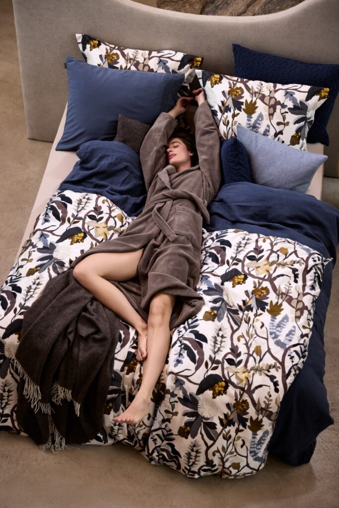 Op een bed met blauw beddengoed met bloemenprint en blauw-witte kussens ligt iemand te ontspannen in een Vandyck Duchess-badjas in de kleur urban taupe, maat XL.