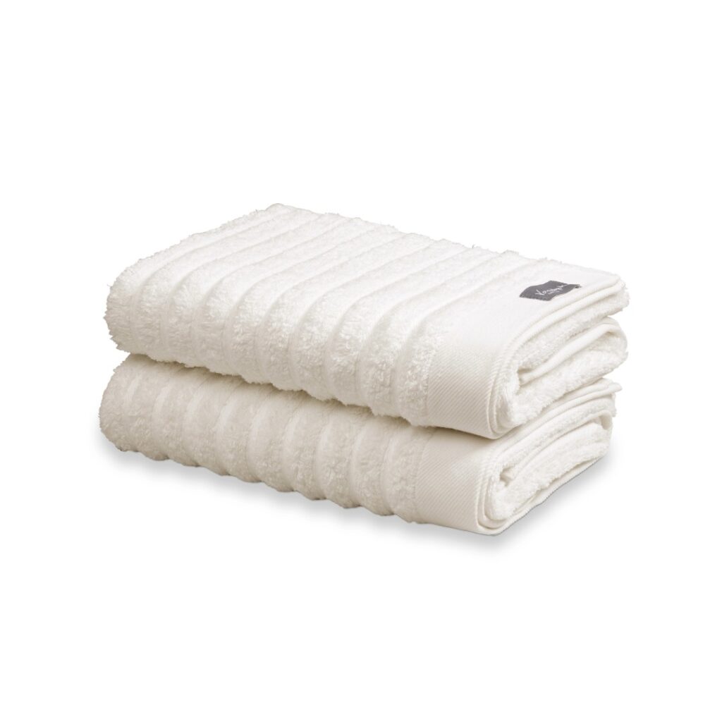 Twee netjes opgevouwen Vandyck Home Wave-gastendoeken in de kleur Cream Tan met geribbelde rand liggen opgestapeld, met zichtbaar merklabel.