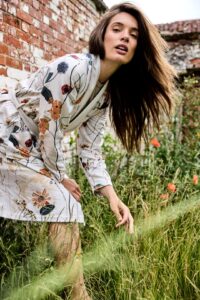 Een vrouw in een Vandyck Nova-jas in de kleur Cream Tan, maat L, buigt voorover in hoog gras met bloemen en bakstenen muren op een zonnige dag.