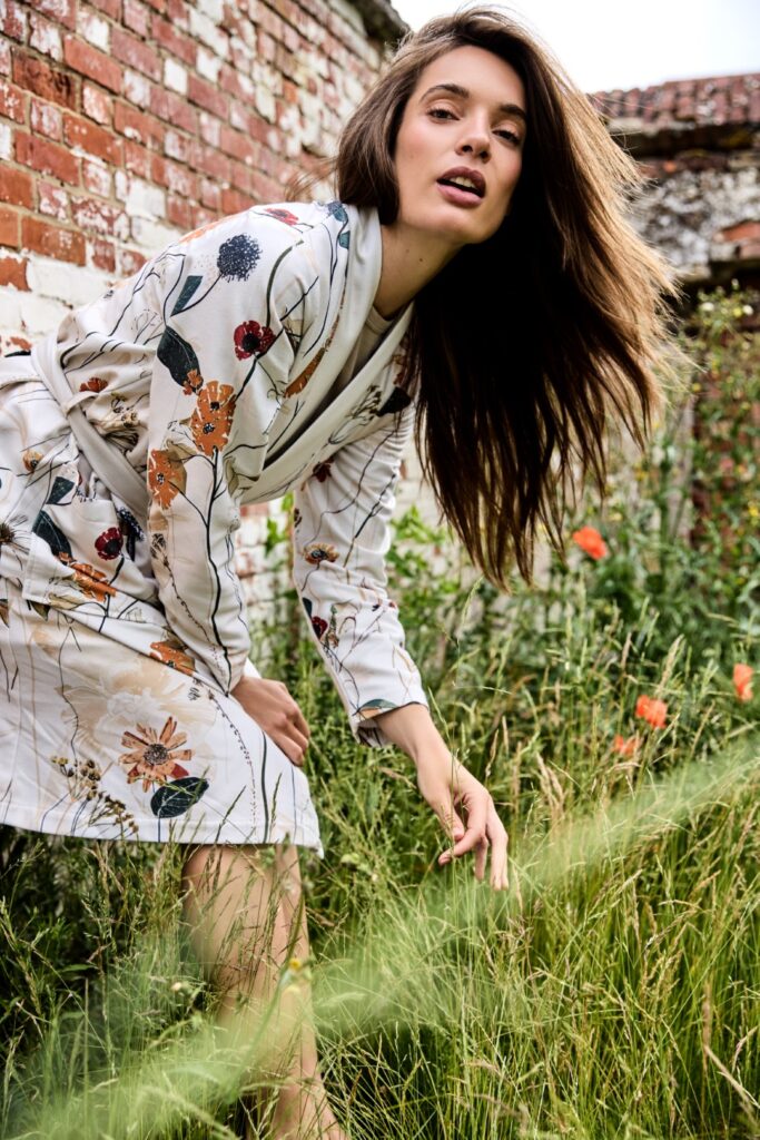 Een vrouw met lang bruin haar straalt zelfvertrouwen uit terwijl ze in de Vandyck Nova-badjas in de kleur Cream Tan XL poseert in een groene tuin bij een verweerde bakstenen muur met wilde bloemen.