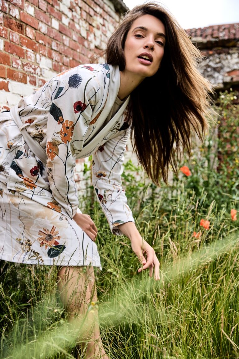 Een vrouw in de Vandyck Nova-badjas in de kleur Cream tan, maat M, leunt voorover in het gras, met rode bakstenen muren en wilde bloemen achter haar.